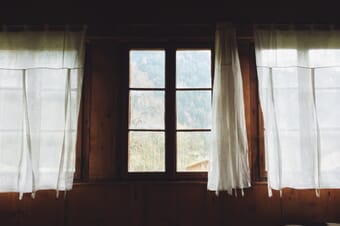 Dark wooden wall with white curtains and a six-panel window showing a forested mountain.