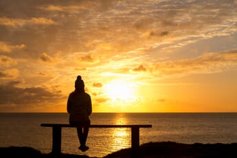 Wide angle shot of the Silhouette of a woman sitting on a bench looking at the tranquil sunset over the ocean.