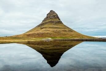 Kirkjufell hill with a clear reflection in the foreground lake, cars on the road at its base, and a moody, overcast sky.