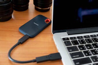 A black external hard drive connected to a MacBook laptop with camera lenses in the background.