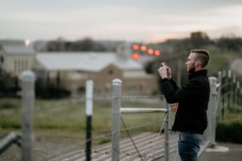 Man taking photo with smartphone by a fence with red bokeh in background.