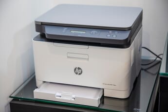 Blue and white HP color laser printer in corner of a room on a glass table with printer tray open