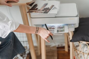 Person's hand pulling printed pages out of a large gray printer while holding a pen.