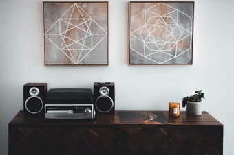 Two art prints with geometric figures hanging on a wall above a table with a sound system, a candle, and a plant.