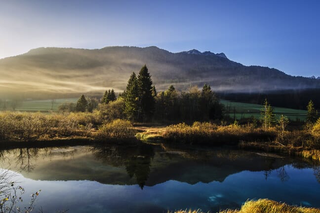 A landscape scene with a pond in the foreground and trees and foggy mountains in the background.