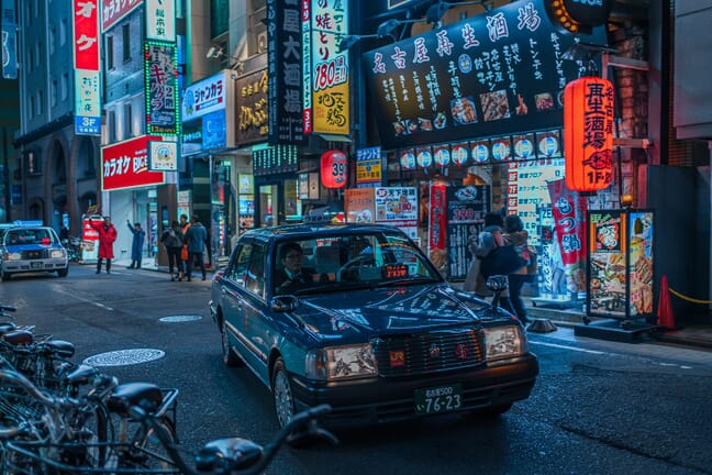 A dimly lit street in Tokyo with a car in the foreground and shoppers and buildings in the background.