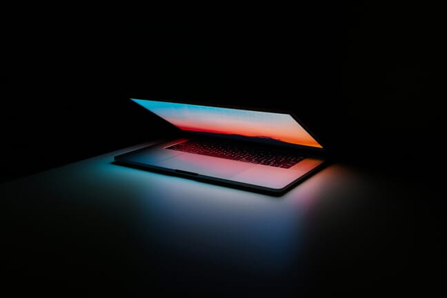 A slightly open laptop with a colorful sunset background in a dark room.
