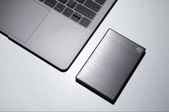 Silver external hard drive on a gray surface next to a gray laptop.
