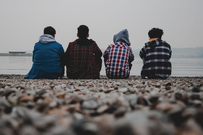 Four teenagers sitting on a beach wearing patterned hoodies and looking out over the water on a gloomy day.