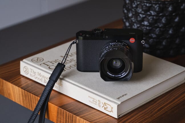 A black Leica camera sits atop a gray book on a light brown coffee table.