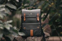 Green Vinta camera backpack with tan leather straps sitting on rocks in a forest surrounded by green leaves.