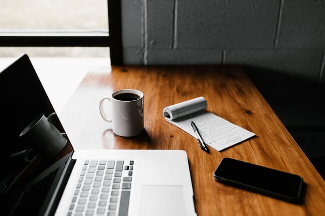 A laptop, a phone, a cup of coffee, and a notepad on a wooden table by a window.