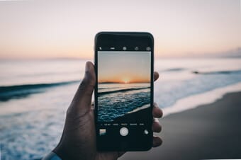 An iphone held up to take a photo of the sunset over a beach.