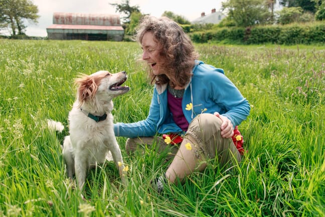 A woman in a blue sweater and her dog sitting in grass looking at each other and laughing.