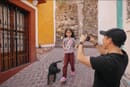 A man wearing a black cap and T-shirt is taking a photo of a young girl with a black dog nearby on a cobblestone street next to brightly painted buildings with wrought iron gates.