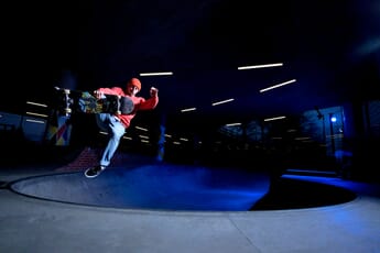 A skateboarder wearing a red beanie and sweatshirt performing a trick in a skatepark bowl at dusk under artificial lights.