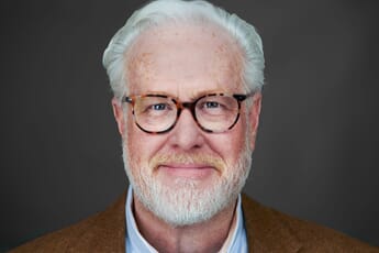 Portrait of a smiling man with white hair and beard, wearing glasses and a brown blazer over a light blue shirt against a dark gray background.