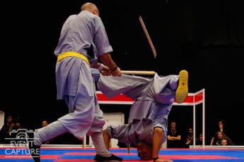 Two men on stage with one man doing a headstand with legs stretched out sideways and the other man hitting him with a stick.