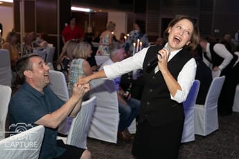 Woman MC at a wedding laughing into microphone as a male guest fake proposes to her.