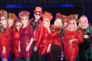 Eight women in red shiny dresses standing four on each side of a man in a red top hat and red shiny jacket.