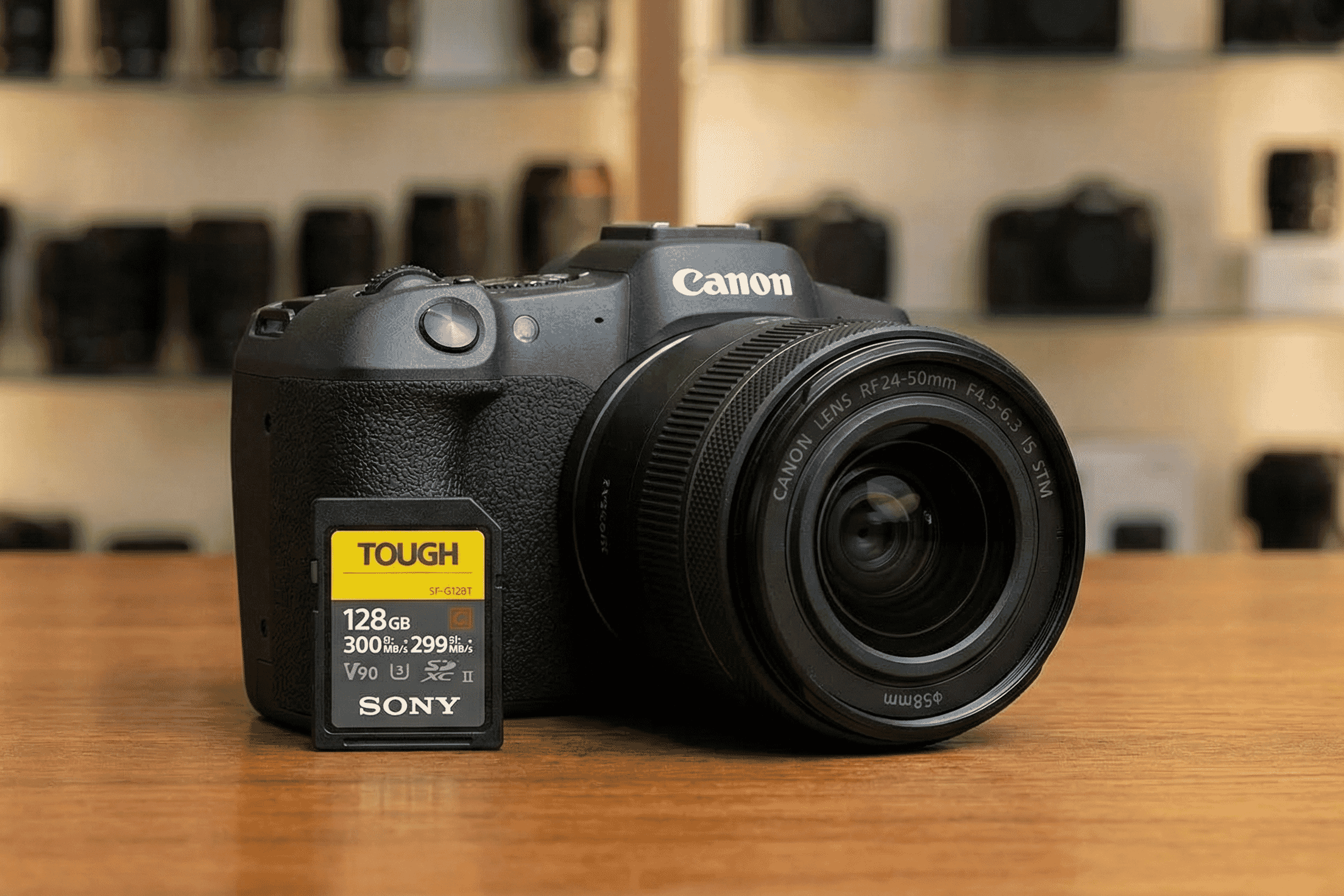 Canon mirrorless camera with RF24-50mm lens and a Sony Tough 128GB SD card displayed on a wooden surface in a camera shop.