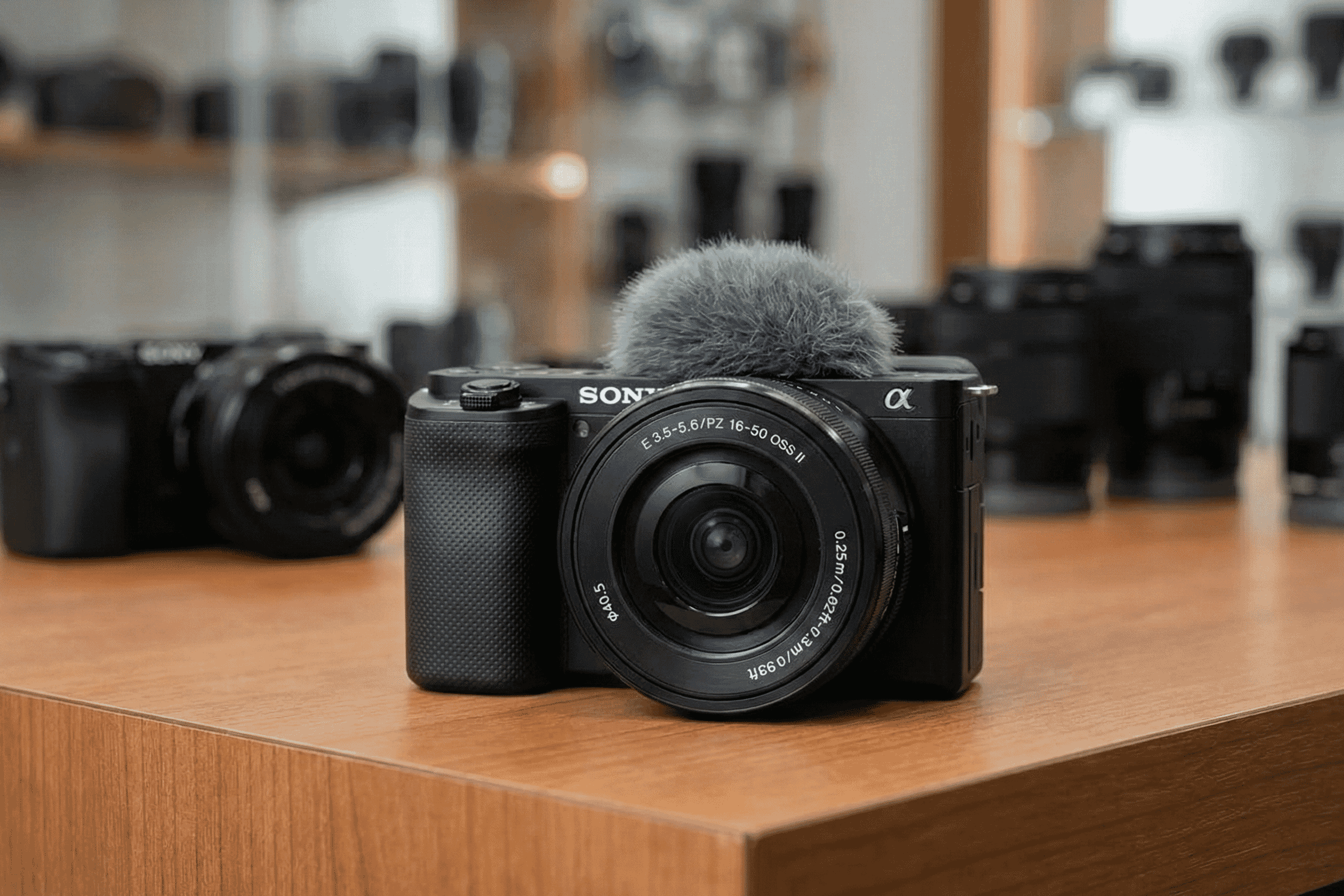 Compact Sony mirrorless vlogging camera with a 16-50mm zoom lens and attached wind muff displayed on a wooden table in a showroom.