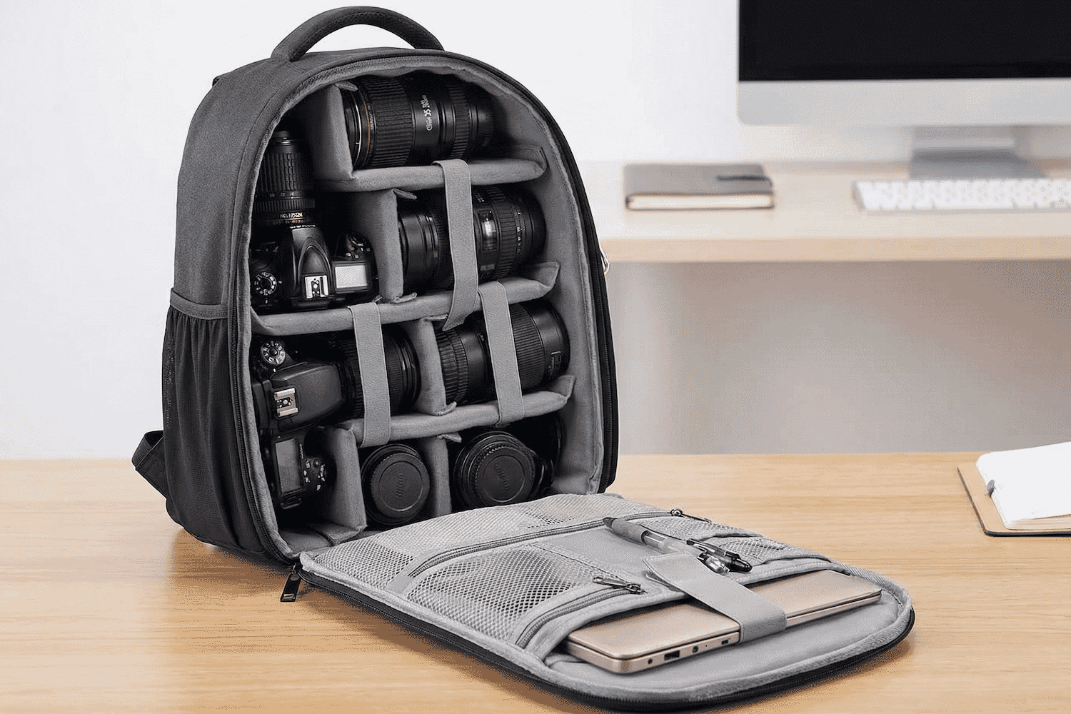 A padded professional camera backpack with organized compartments holding DSLR cameras, multiple lenses, and accessories on a wooden desk near a computer.