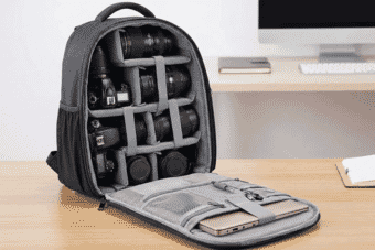 A padded professional camera backpack with organized compartments holding DSLR cameras, multiple lenses, and accessories on a wooden desk near a computer.