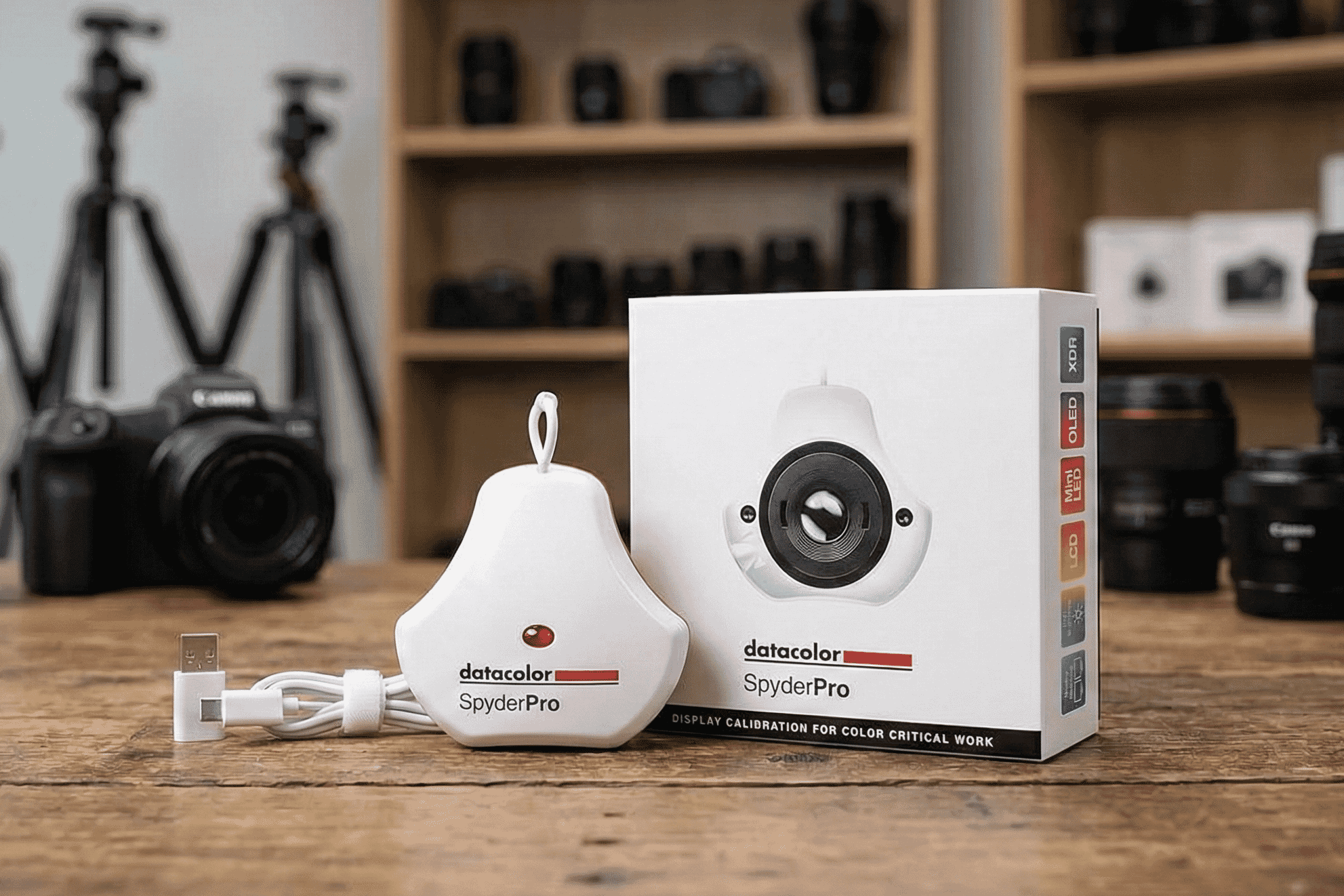 The Datacolor SpyderPro display calibration device sits beside its colorful retail box on a wooden desk in a photography studio setup.