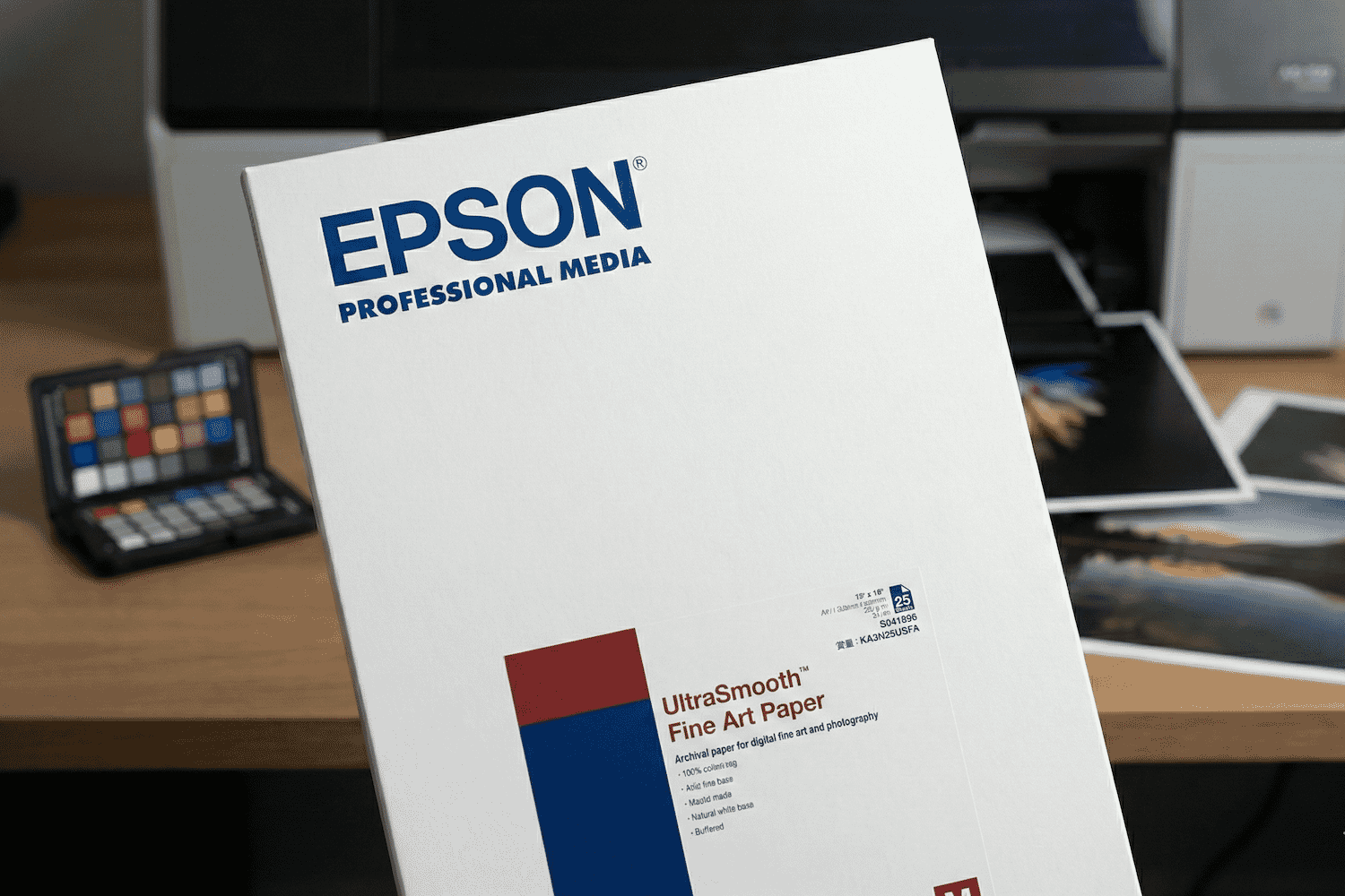 Epson UltraSmooth Fine Art Paper box displayed on a desk with printed photos and a color calibration chart in the background.