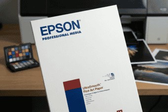 Epson UltraSmooth Fine Art Paper box displayed on a desk with printed photos and a color calibration chart in the background.