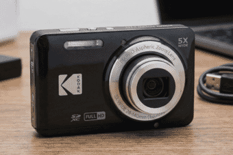 A compact black Kodak PixPro digital camera with a 5X zoom lens sits on a wooden desk near a USB cable and laptop.