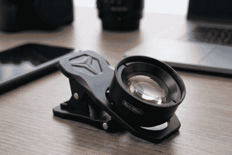 A high-quality 100mm macro smartphone lens clip rests on a modern wooden desk, surrounded by photography gear and sleek tech accessories.