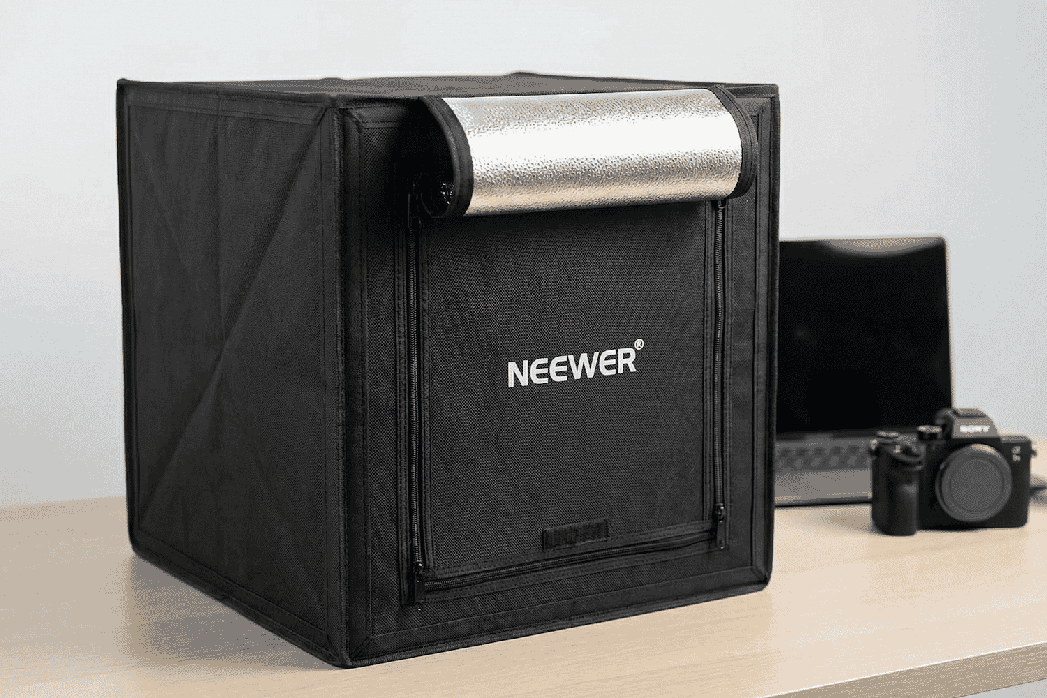 A portable Neewer photo light box with a reflective silver interior is set up on a desk beside a camera and laptop, ideal for product photography.