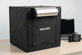 A portable Neewer photo light box with a reflective silver interior is set up on a desk beside a camera and laptop, ideal for product photography.