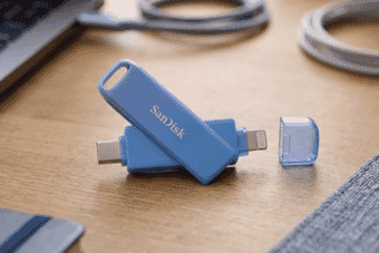 A sleek blue SanDisk dual USB flash drive with USB-C and Lightning connectors rests on a wooden desk near a laptop and cables, offering portable data storage.