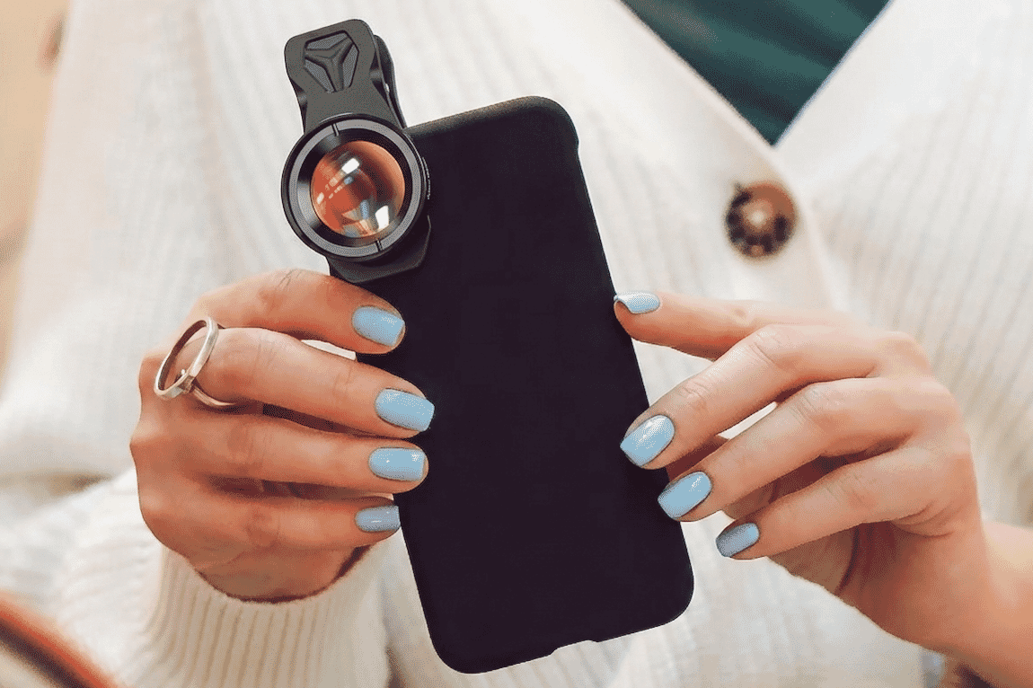 A woman with pastel blue nails holds a smartphone with a detachable macro lens, showcasing mobile photography accessories for content creators.
