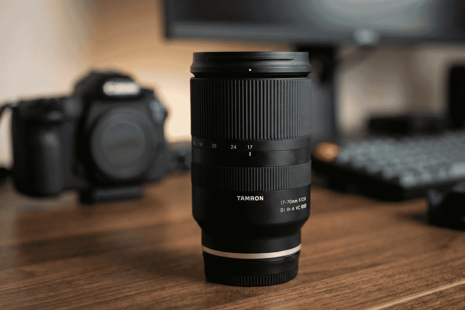A Tamron 17-70mm f/2.8 Di III-A VC RXD lens stands upright on a wooden desk beside a blurred DSLR camera and keyboard in the background.