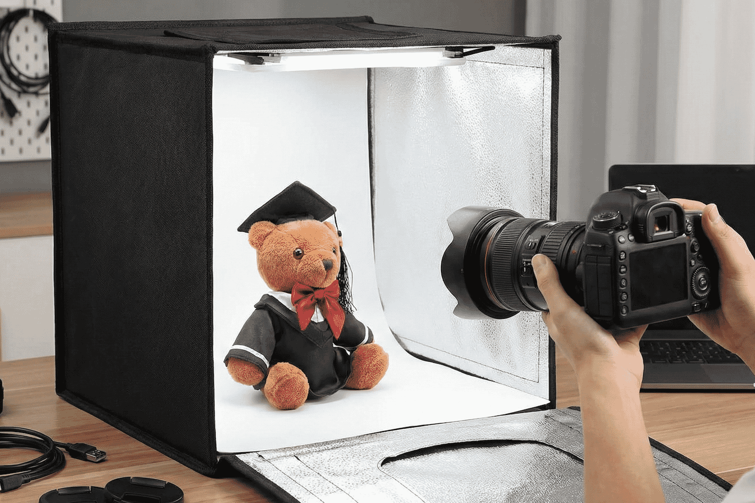 A soft, brown teddy bear in a graduation outfit is being photographed in a portable light box studio using a professional DSLR camera.