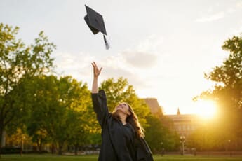 A graduate in cap and gown tossing their cap at golden hour on campus